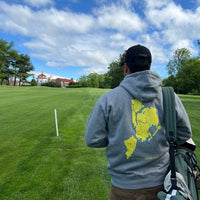 Person wearing a gray sweatshirt with a map design on a grassy field with trees and a blue sky.