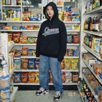 Female model wearing a black hoodie with 'Queens' text in a store aisle with snacks and drinks. QCC Varsity Script Hoodie