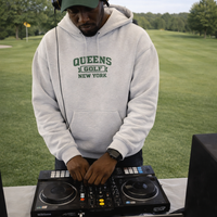 Person wearing a 'Queens Golf New York' hoodie and cap, standing behind DJ equipment on a grassy area.