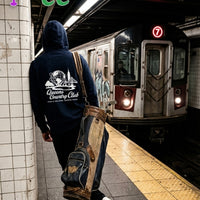Person in a hoodie with 'Queens Country Club' logo at a subway station, train visible.