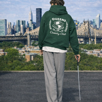 Model with Queens Unisphere Golf Ball Tee Hoodie Forest Green Back looking at NYC skyline on the rooftop in Queens
