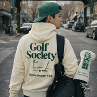 Person wearing a hoodie with 'Golf Society' text, walking on a street in Queens.