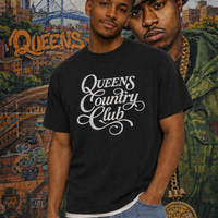 Model wearing a black QCC Heritage Script heavyweight tee in front of a Queens mural