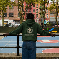 Person wearing a green hoodie with a queens borough golf recreation logo and text, standing on a basketball court with trees and buildings in the background.