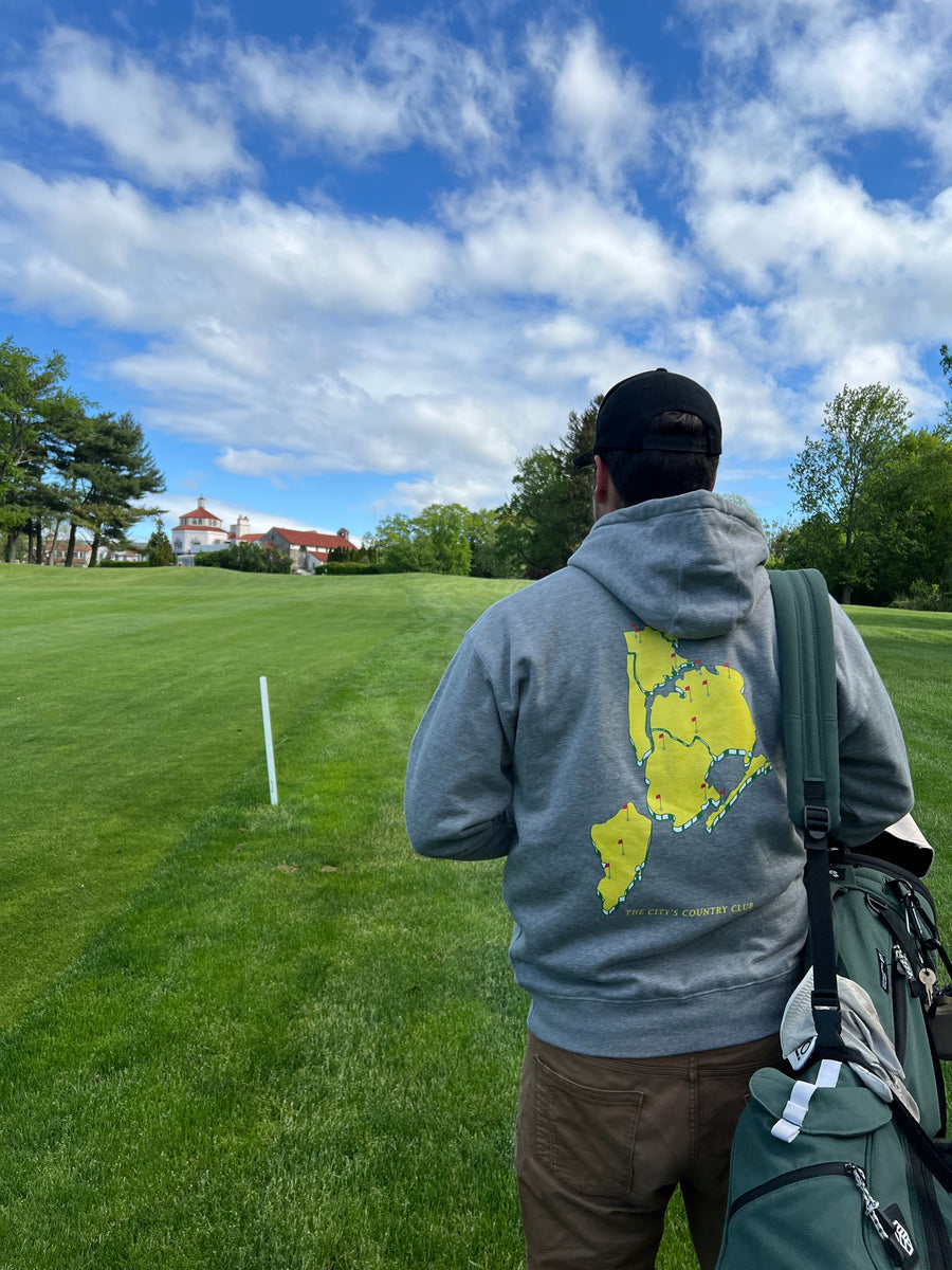 Person wearing a gray sweatshirt with a map design on a grassy field with trees and a blue sky.
