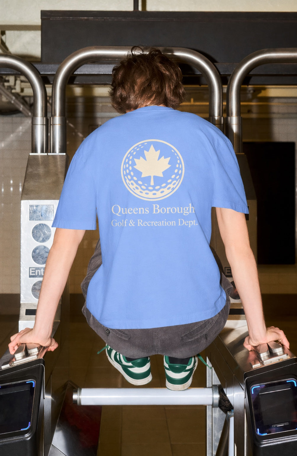 Person wearing a blue t-shirt with a Queens Borough Golf & Recreation logo and text, standing at a security checkpoint.