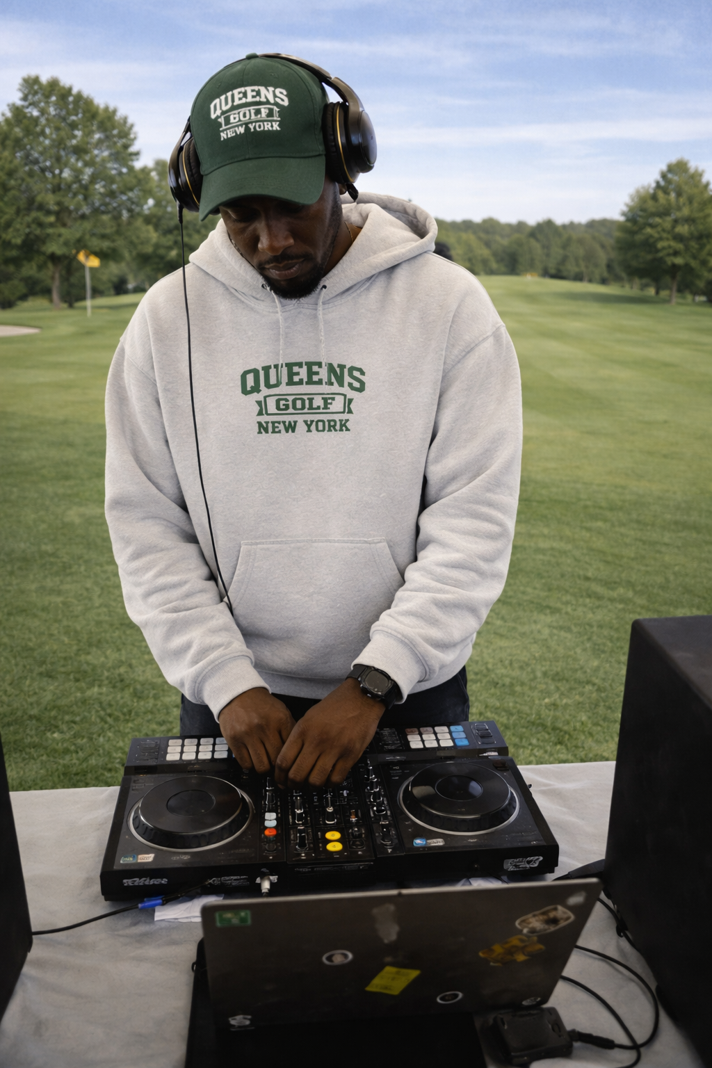 Person wearing a 'Queens Golf New York' hoodie and cap, standing behind DJ equipment on a grassy area.