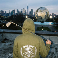 Model wearing QCC golf ball tee hoodie looking at nyc skyline and queens unisphere 