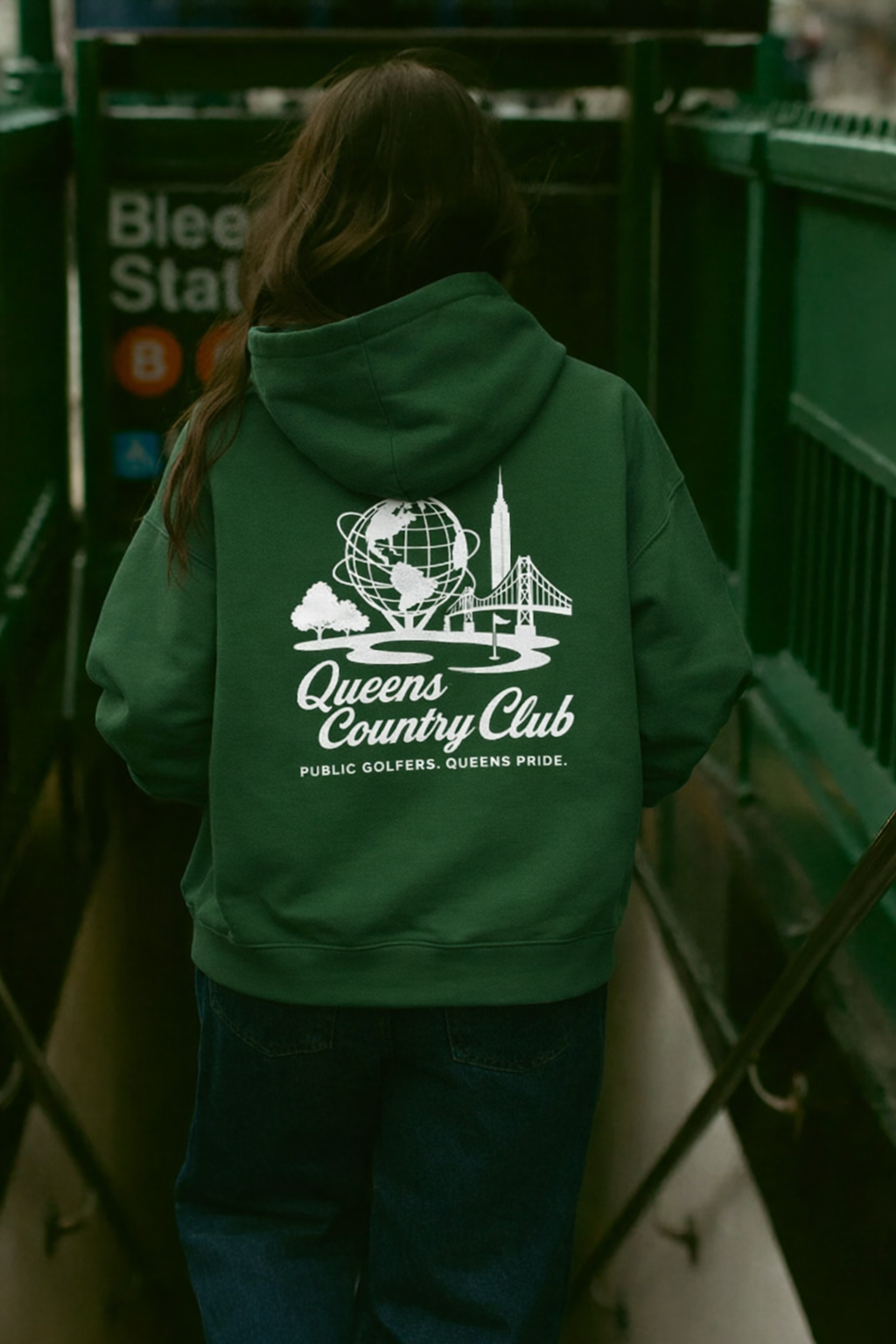 Person wearing a green hoodie with 'Queens Country Club' logo in an urban setting.