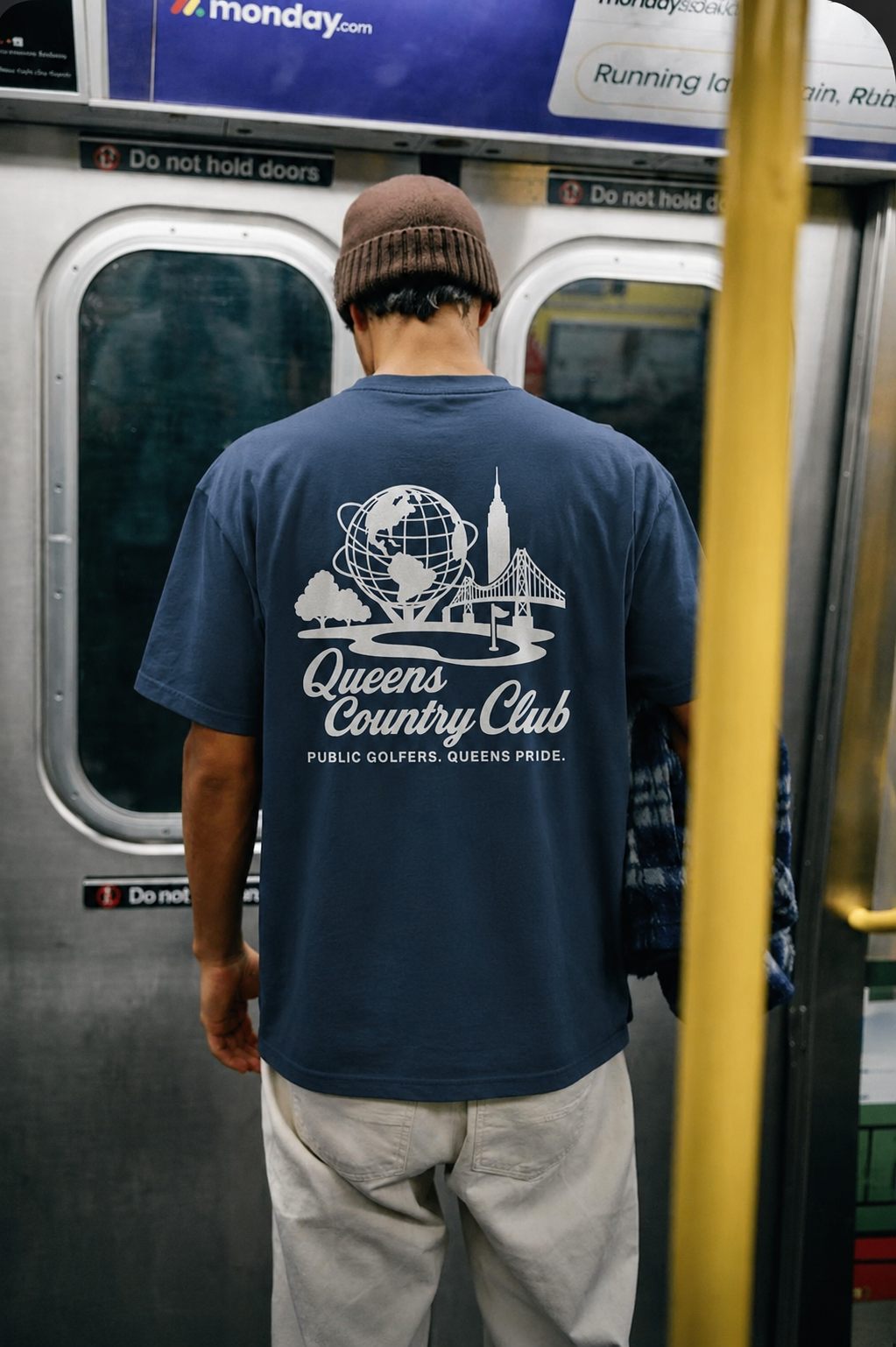 Person wearing a blue t-shirt with 'Queens Country Club' design on a subway train.