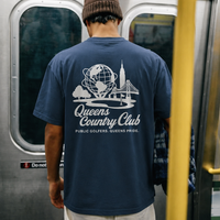 Person wearing a blue t-shirt with 'Queens Country Club' design on a subway train.