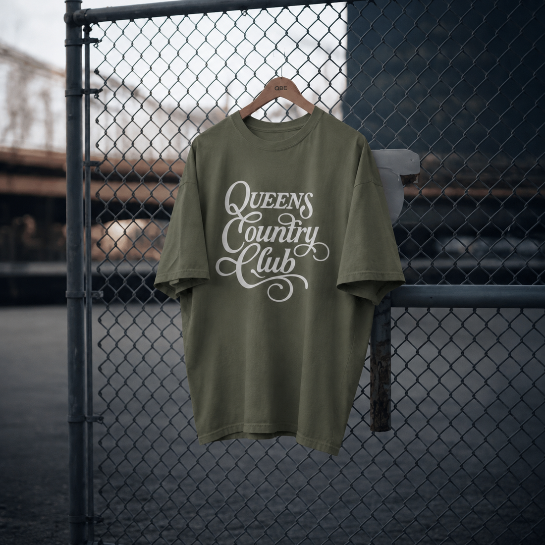 Moss QCC Heritage Script heavyweight tee displayed on a hanger against a chain-link fence