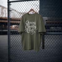 Moss QCC Heritage Script heavyweight tee displayed on a hanger against a chain-link fence