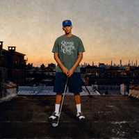 Golfer wearing a moss QCC Heritage Script heavyweight tee on a Queens rooftop at sunset