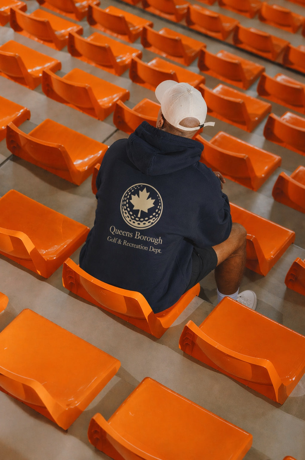 Person wearing a hoodie with a queens borough golf & recreation logo sitting among orange chairs in sports stadium