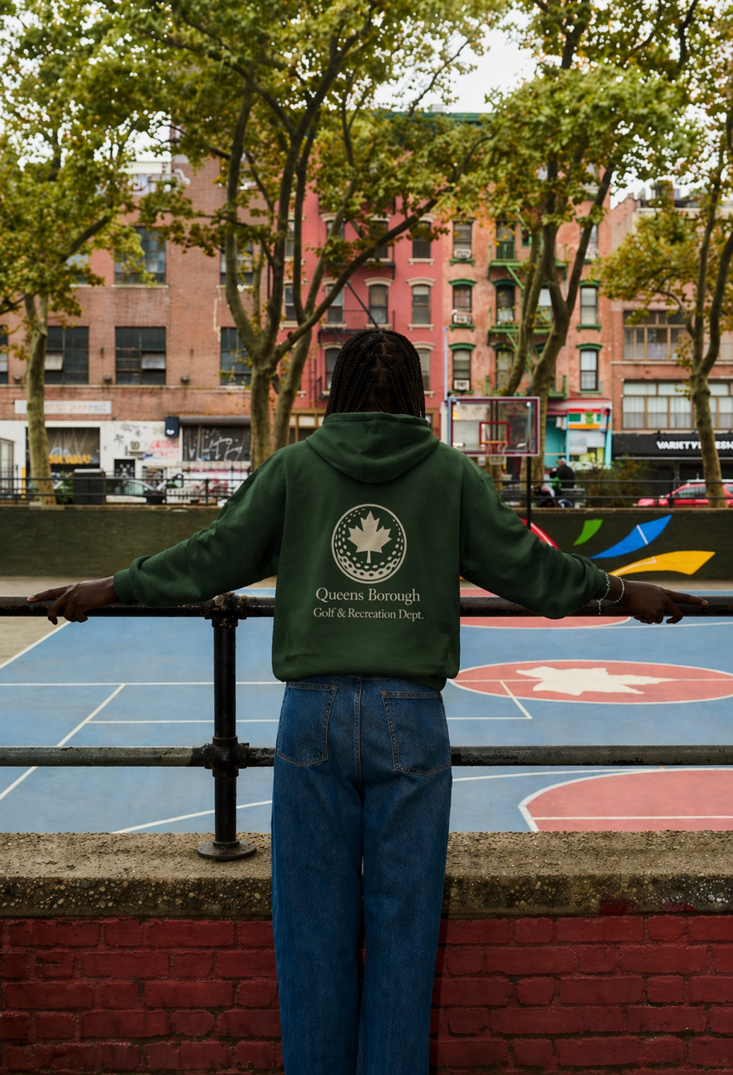 Person wearing a green hoodie with a queens borough golf recreation logo and text, standing on a basketball court with trees and buildings in the background.